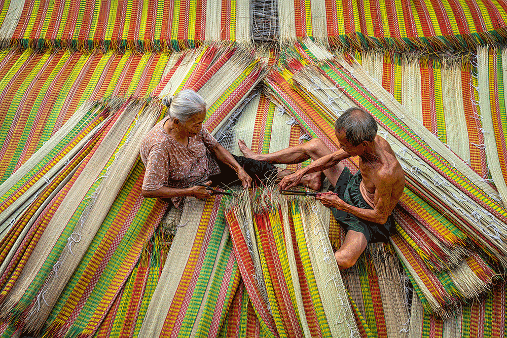A top-down perspective showcasing the intricate, handcrafted process of a Vietnamese artisan creating a woven mat on Cam Kim Island, Hoi An.
